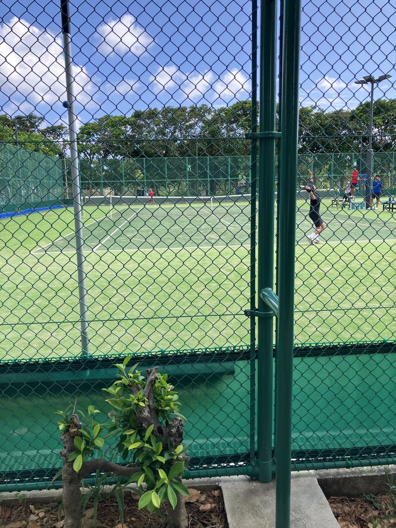 フェンス越しに見たテニスの試合会場。青空の下、コートでサーブを打つ選手の姿